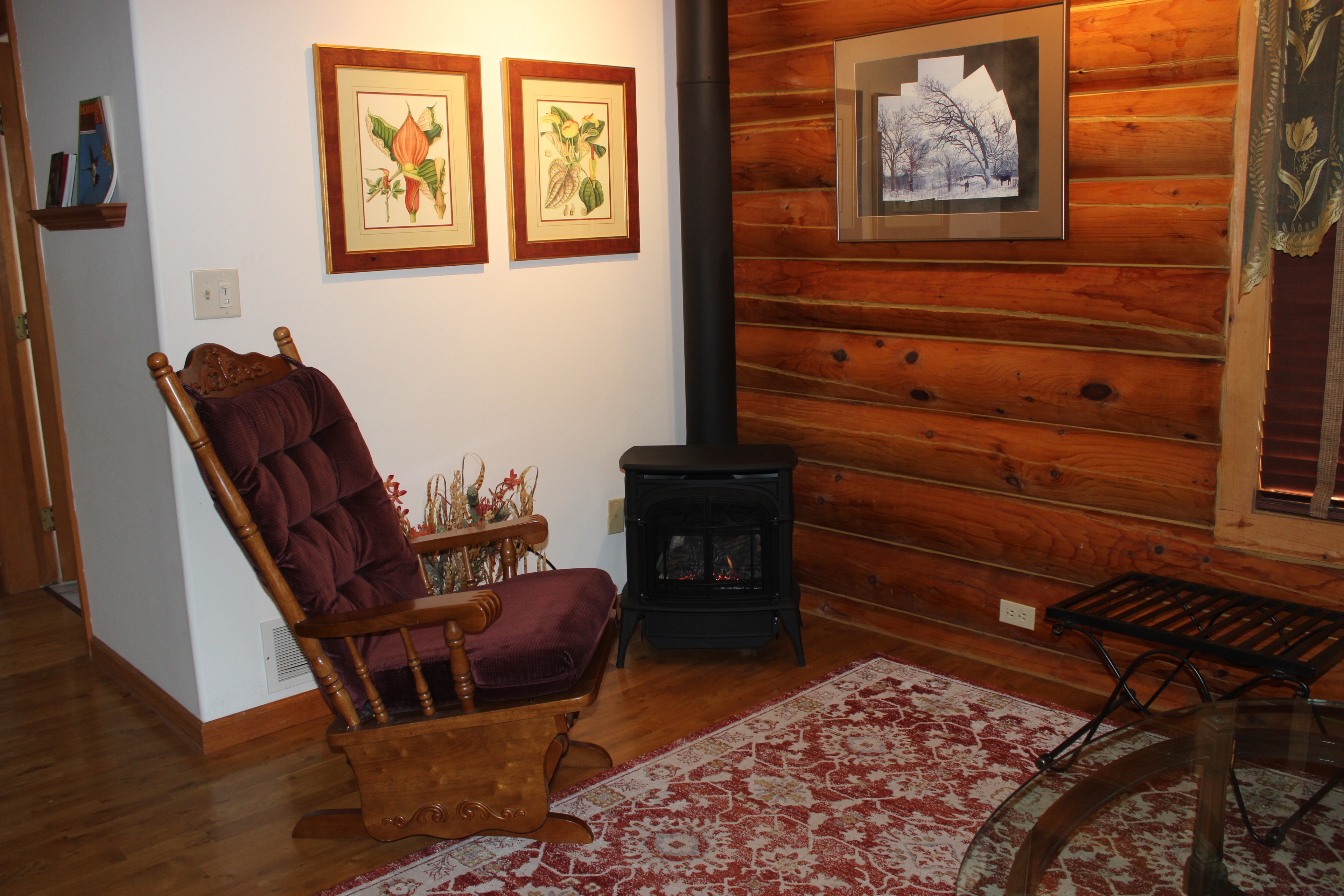 Sitting area in a log cabin room with wooden glider rocker, red and cream patterned area rug, and fireplace.