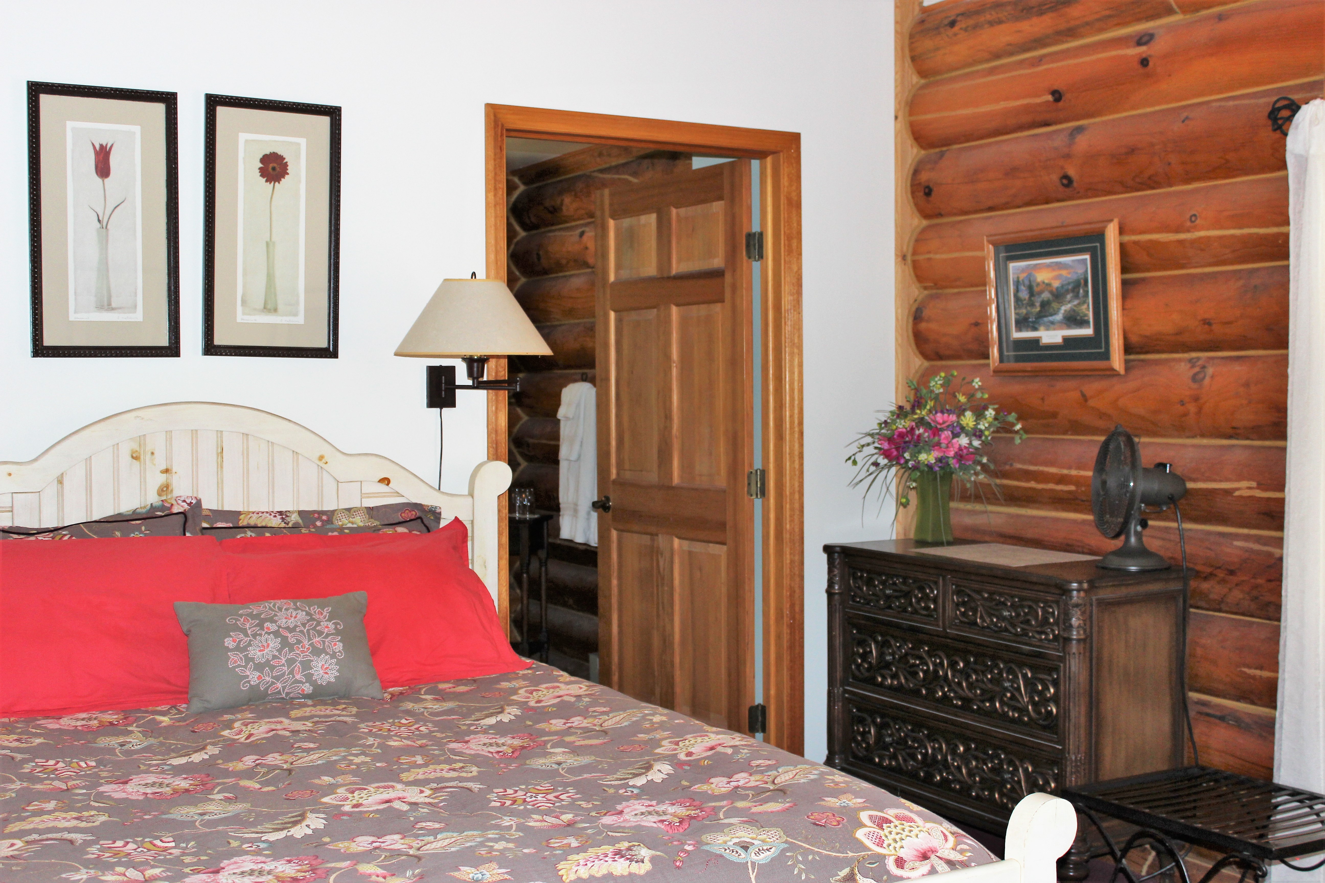 Cozy log cabin bedroom featuring a bed with a floral comforter and bright red pillows, an ornate dark wood dresser with a small fan and flower vase, and rustic log walls. The room includes framed floral artwork above the bed and an open wooden door leading to private bathroom.