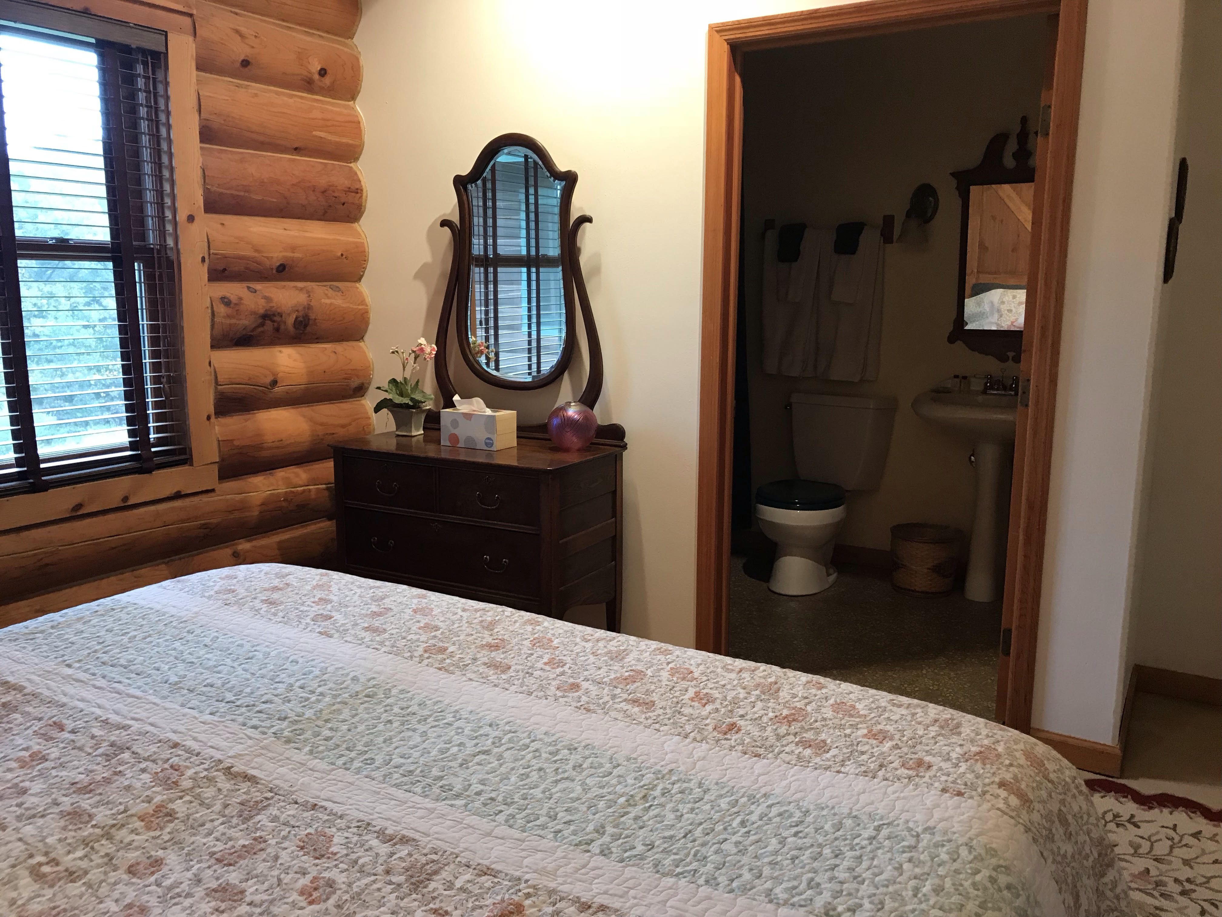A rustic bedroom inside a log cabin featuring a quilted bed in the foreground and a dark wood dresser topped with a decorative oval mirror. The room has a large window set into a log-timber wall. An open doorway provides a clear view into an en-suite bathroom containing a white pedestal sink and a toilet.