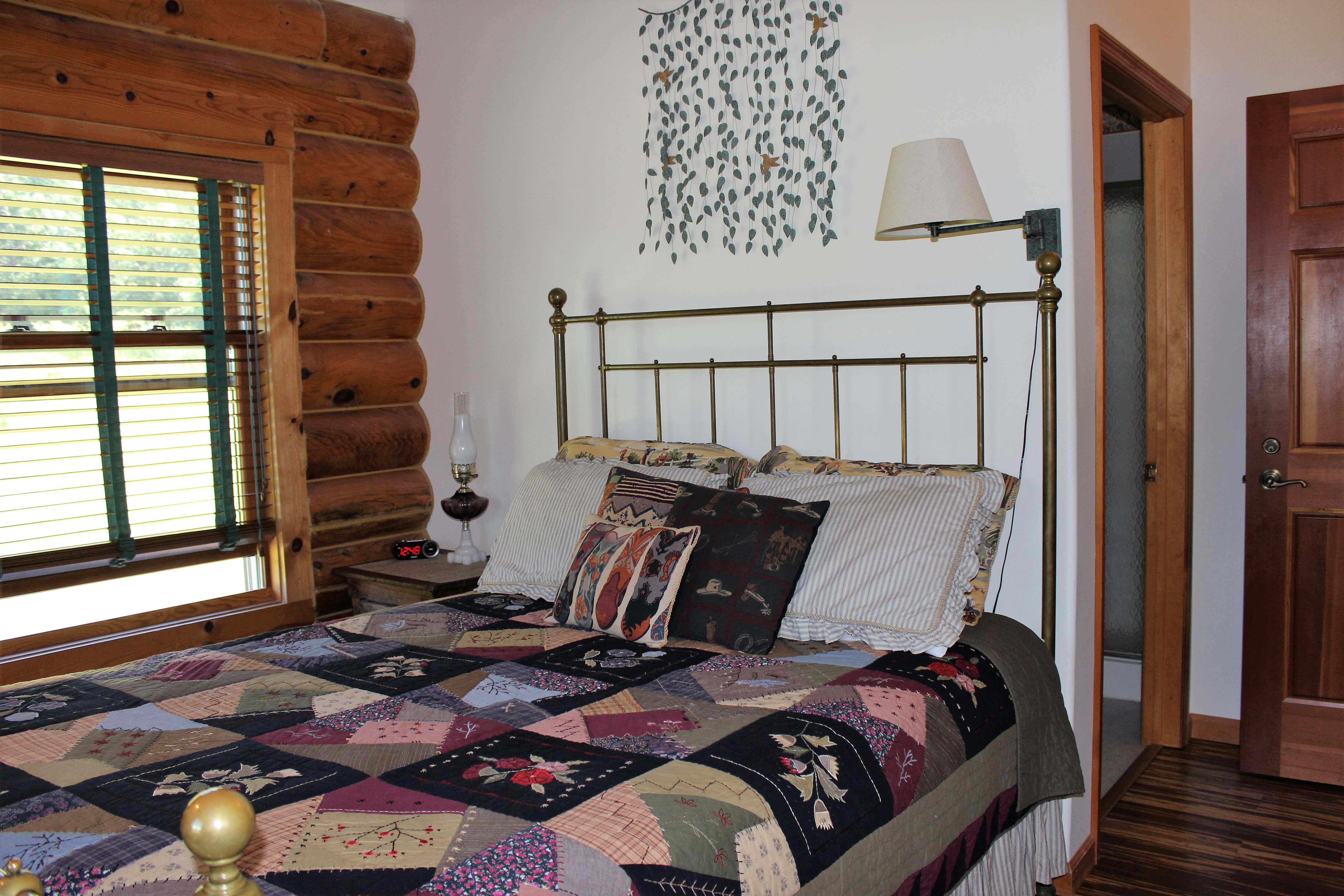 A cozy cabin bedroom featuring a brass bed frame with a patchwork quilt in the foreground. The room has a large window with horizontal blinds set into a log-timber wall. To the right of the bed, a white wall is decorated with a metal wall hanging, and an open doorway leads to a bright bathroom.