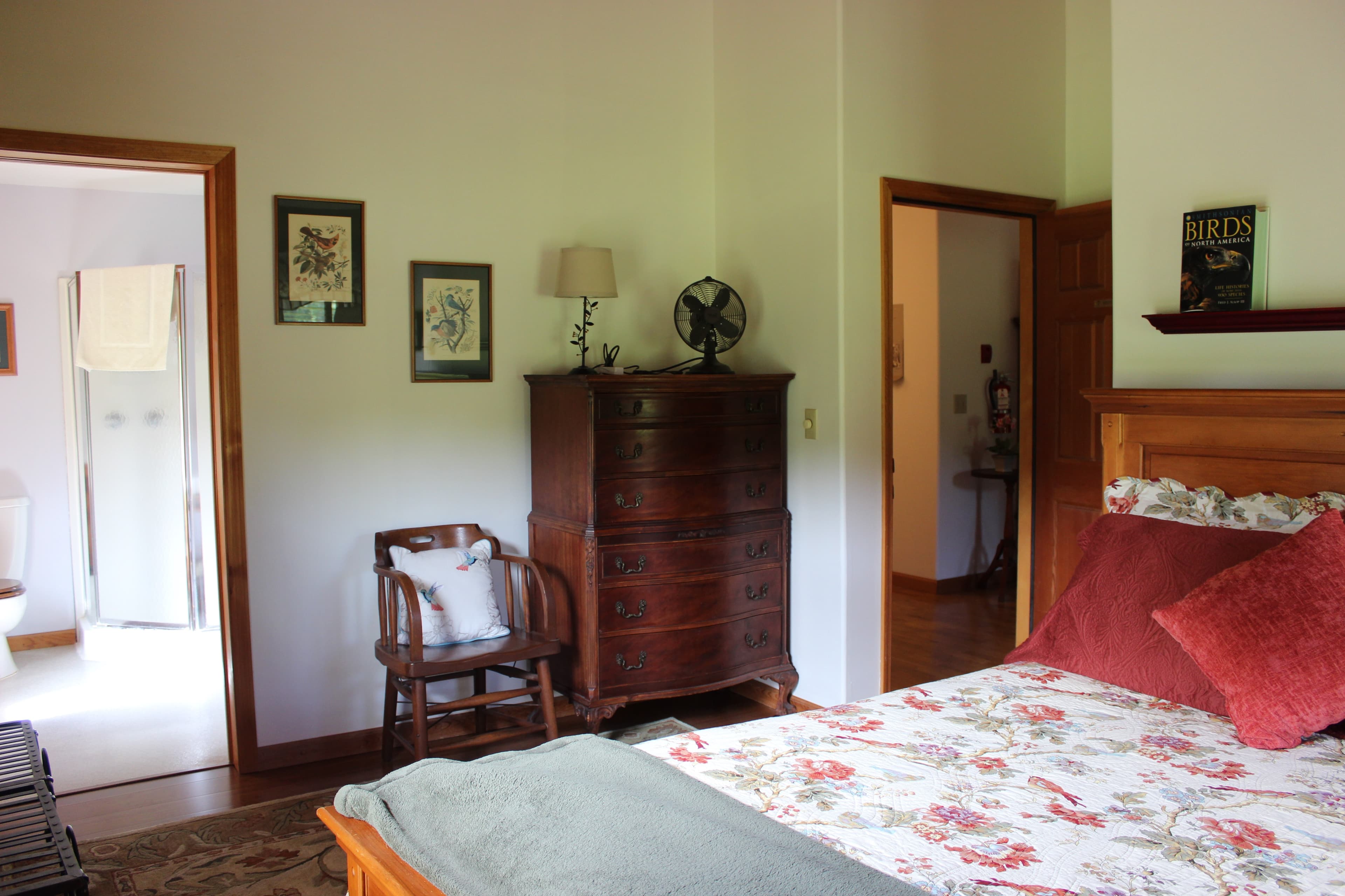 A cozy bedroom featuring a floral-patterned bed with vibrant red pillows and a green throw blanket in the foreground. Against the light green walls sits a tall, dark wood dresser with a small vintage-style fan and a lamp on top. To the left, an open doorway reveals a bright, white-tiled bathroom, while another open door to the right shows a glimpse of a hallway.