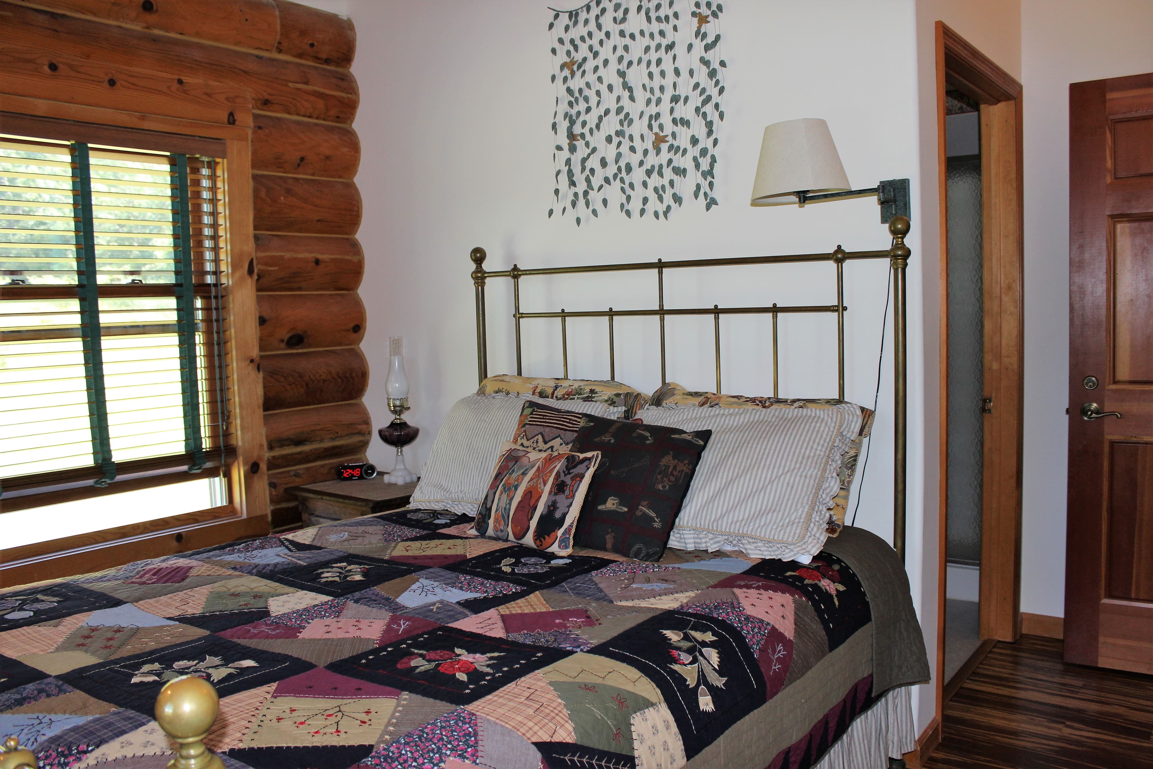 A cozy cabin bedroom featuring a brass bed frame with a patchwork quilt in the foreground. The room has a large window with horizontal blinds set into a log-timber wall. To the right of the bed, a white wall is decorated with a metal wall hanging, and an open doorway leads to a bright bathroom.
