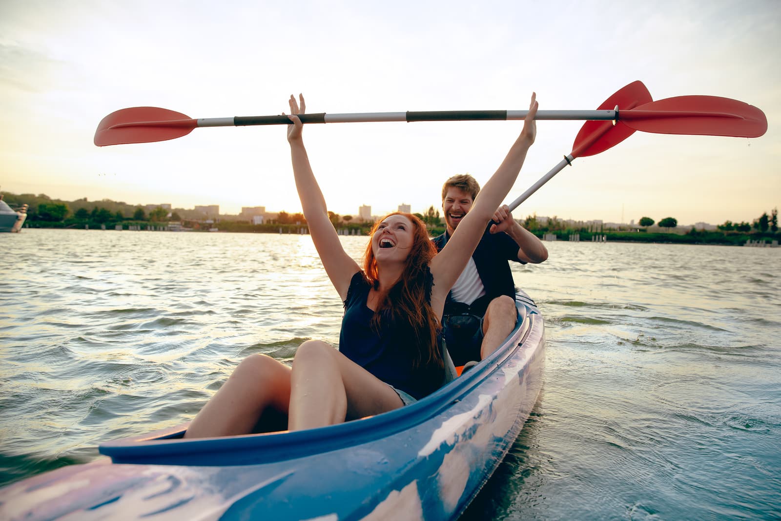 A man and woman joyfully kayak on the water, with the woman raising a paddle in celebration.