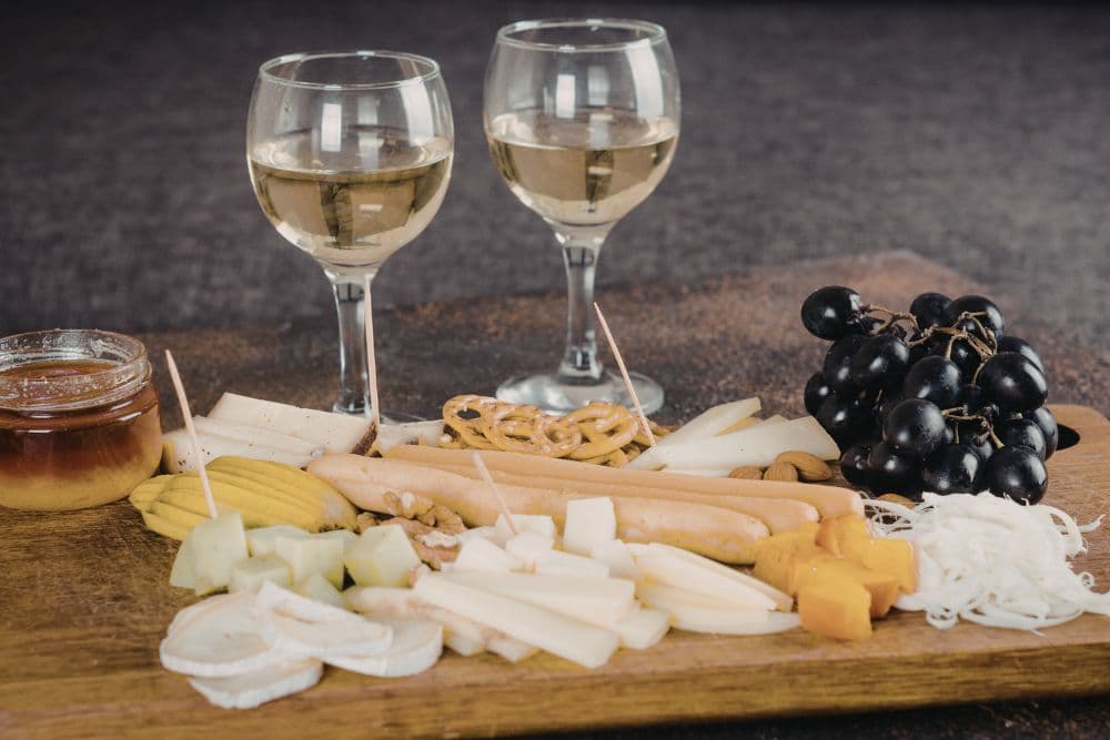 A charcuterie board with various cheeses, fruits, pretzels, and honey, accompanied by two glasses of white wine.