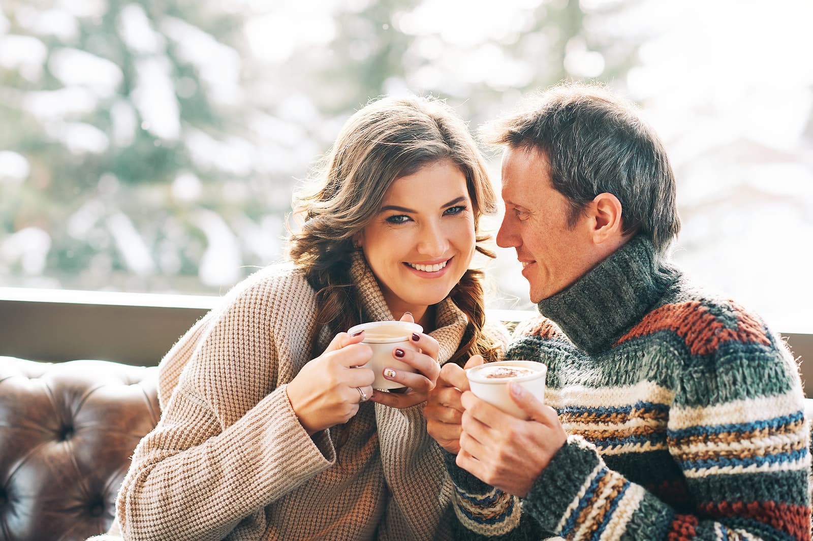 A couple sits together on a cozy sofa, smiling and holding warm mugs in a winter setting. A couple sits together on a cozy sofa, smiling and holding warm mugs in a winter setting.