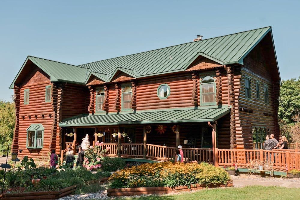 A large log cabin with green roofing surrounded by gardens and people.