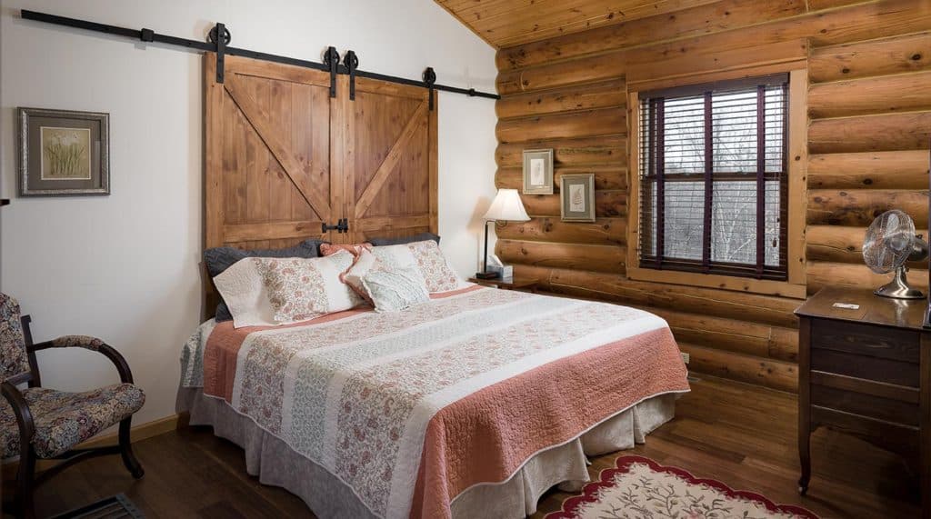 A cozy bedroom with a wooden log wall, featuring a large bed with a patterned quilt, a bedside table, and a chair.