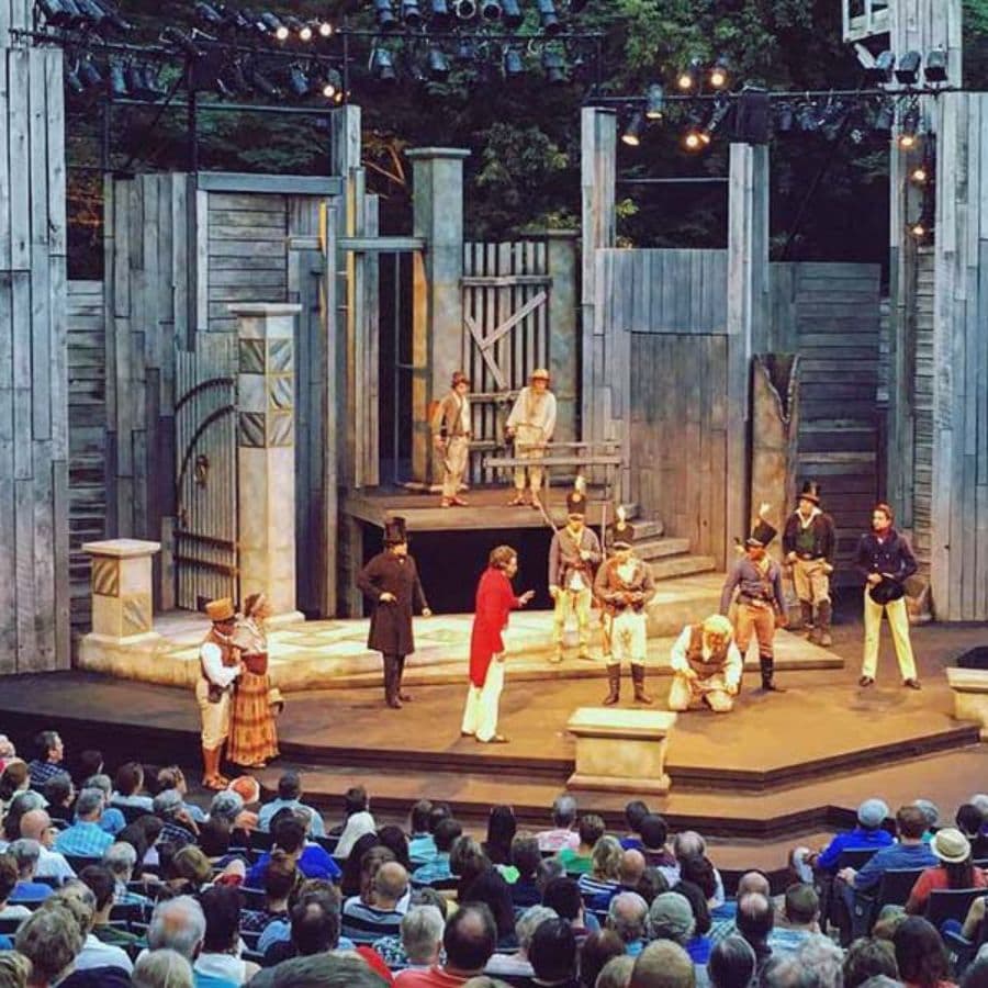 A group of actors performs on stage in front of an audience in an outdoor theater.