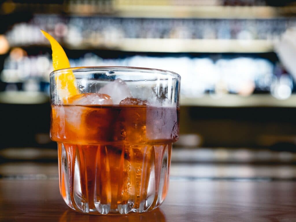 A glass of cocktail with ice and a lemon twist, set against a blurred bar background.