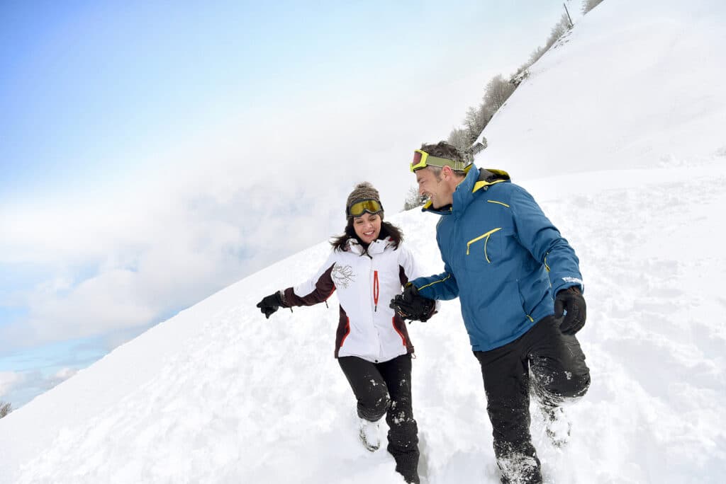 Two people joyfully running through deep snow against a backdrop of a blue sky.