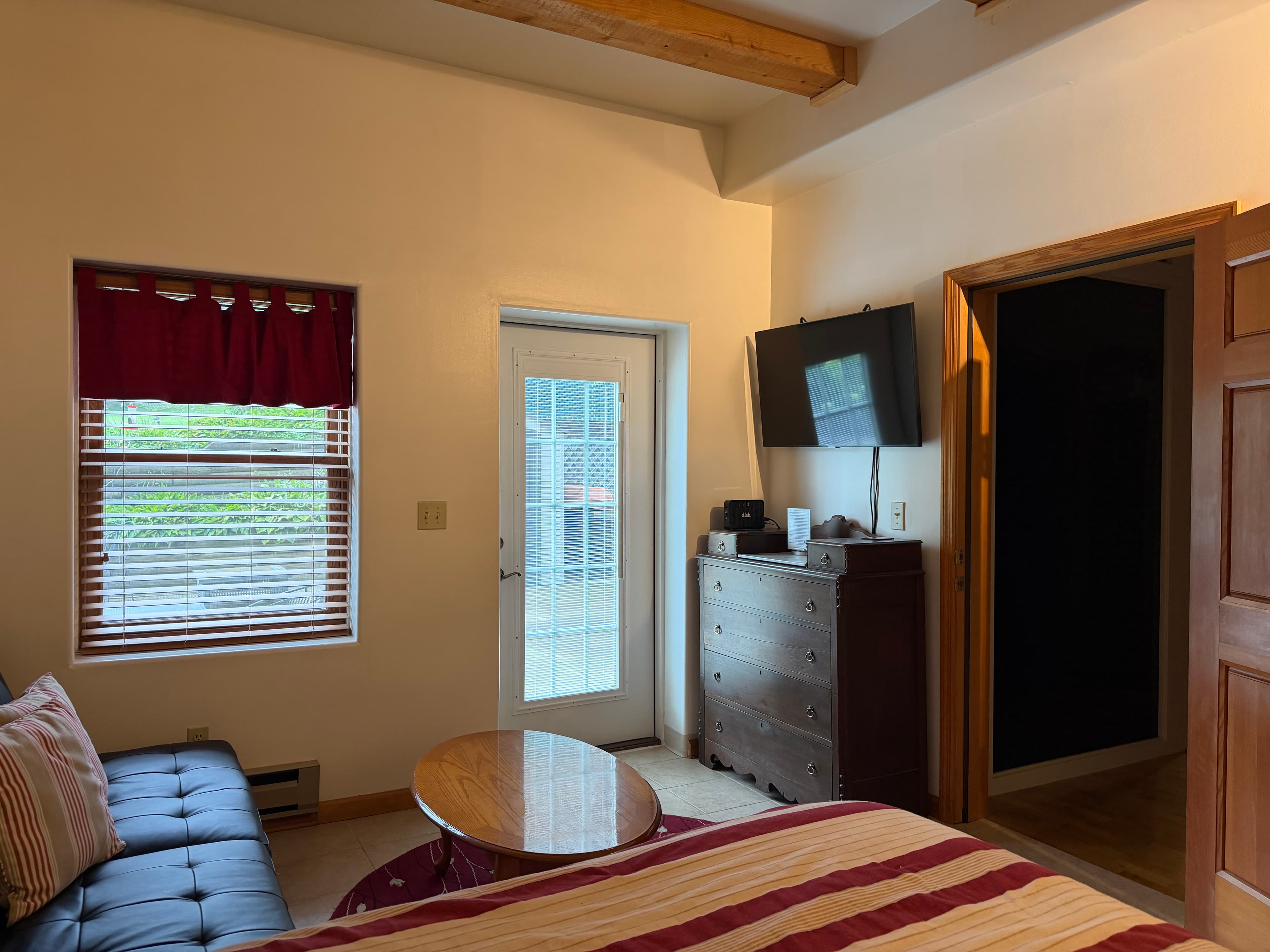 A cozy bedroom featuring a bed, couch, window with blinds, wooden dresser, and a door leading outside.