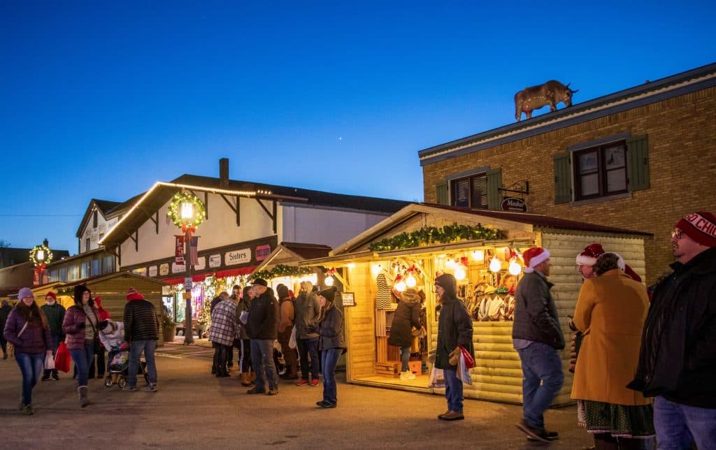 A festive market scene with people shopping at holiday stalls under a twilight sky, featuring buildings adorned with lights.