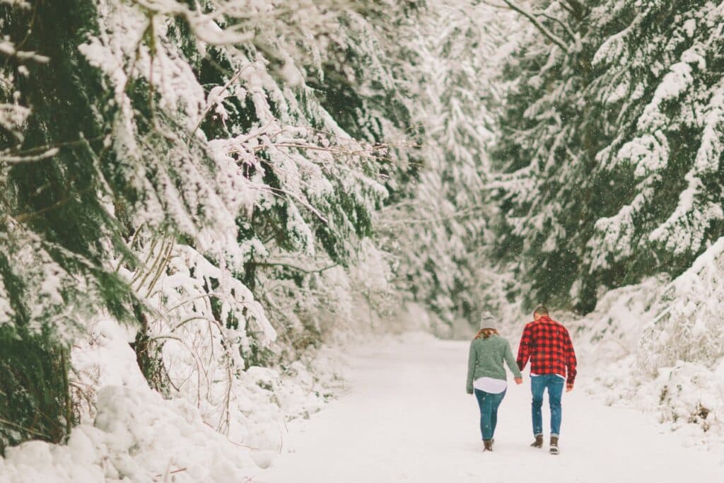 A couple walks hand-in-hand down a snow-covered path surrounded by evergreen trees.