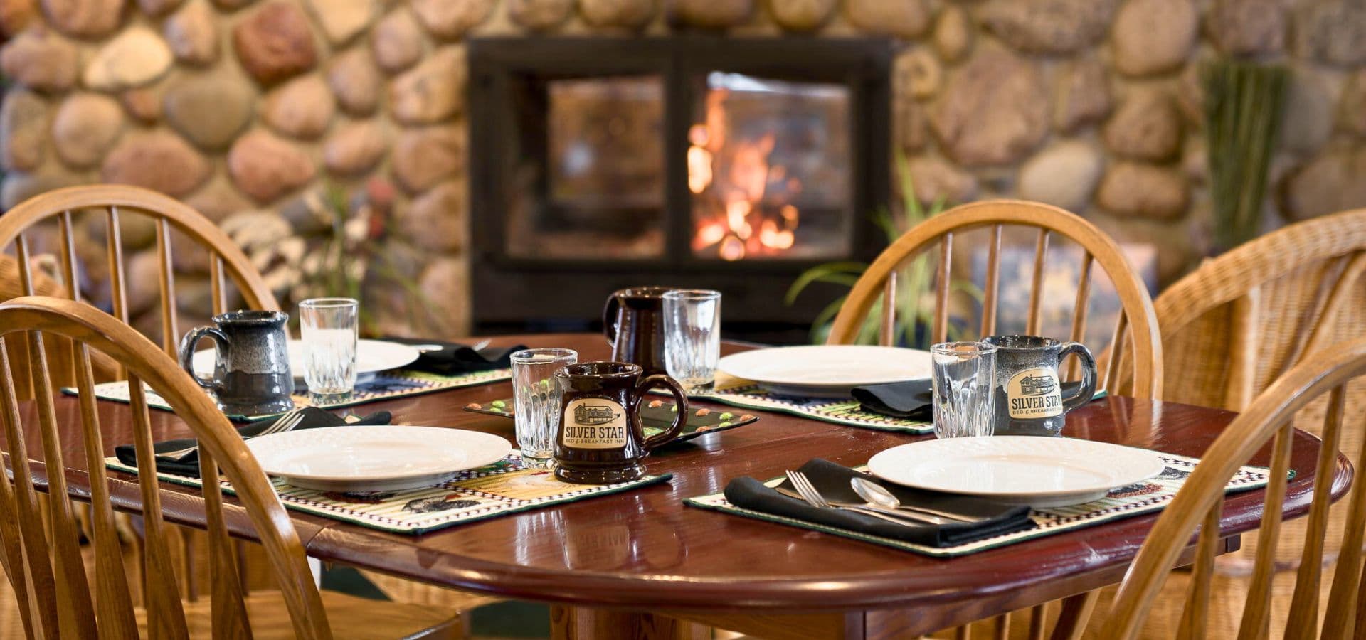 A wooden dining table set with plates and glasses, in front of a cozy stone fireplace.