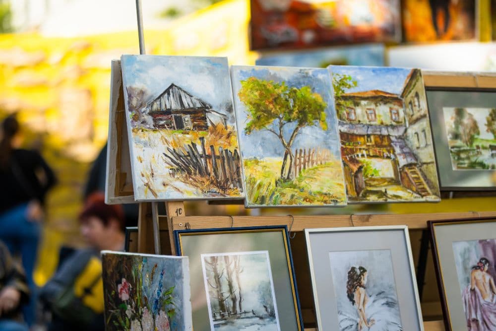 A display of various colorful paintings, including landscapes and a figure, at an outdoor art fair.