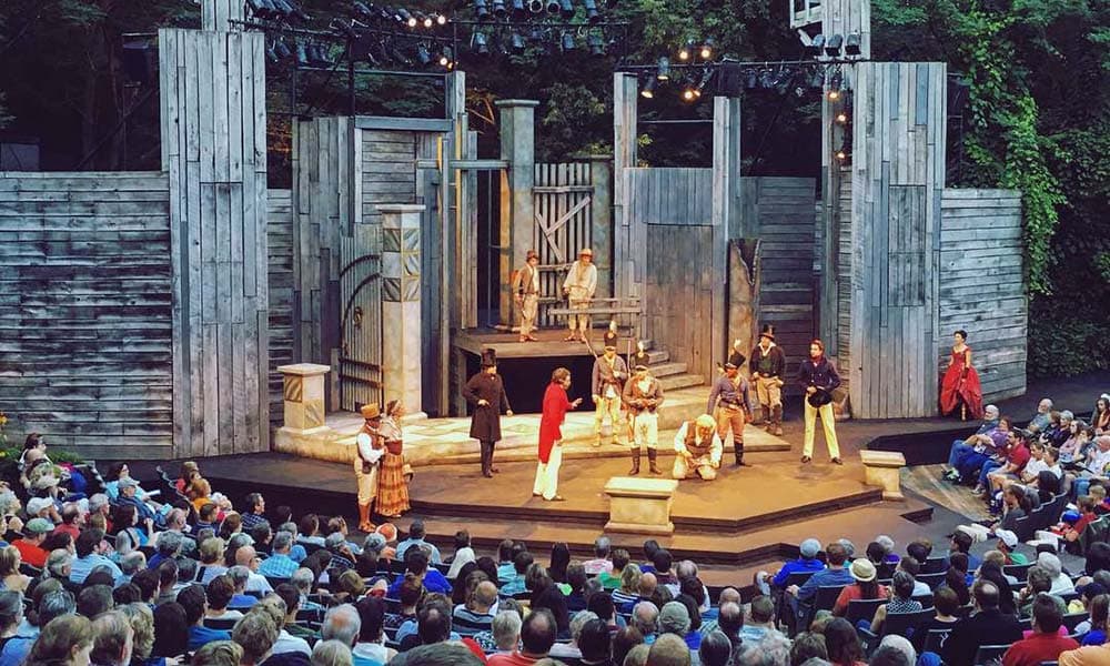 A theater performance taking place outdoors with an audience seated in front of a wooden stage.