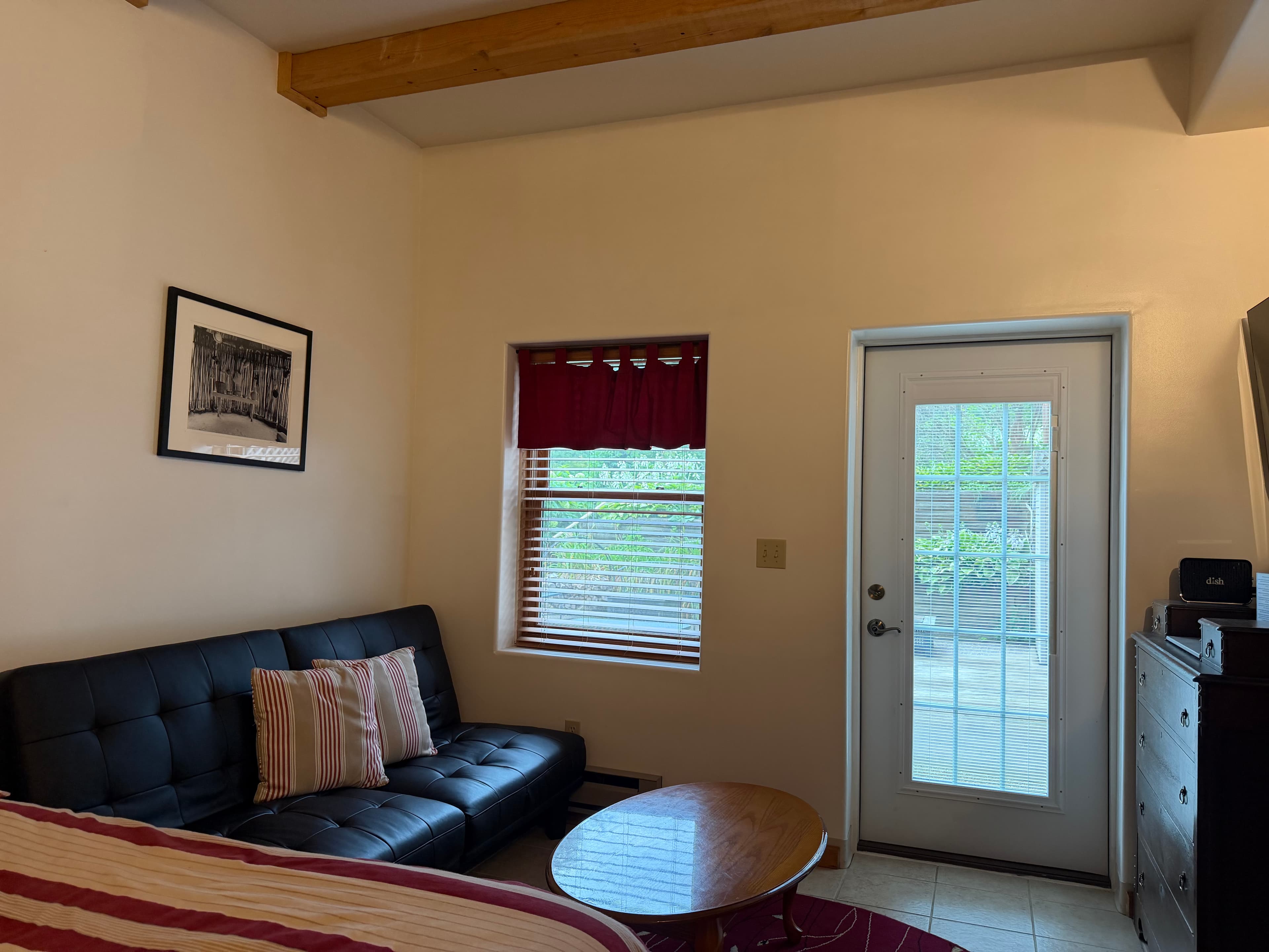A cozy interior featuring a black couch, a round wooden coffee table, a window with red curtains, and a glass door leading outside.